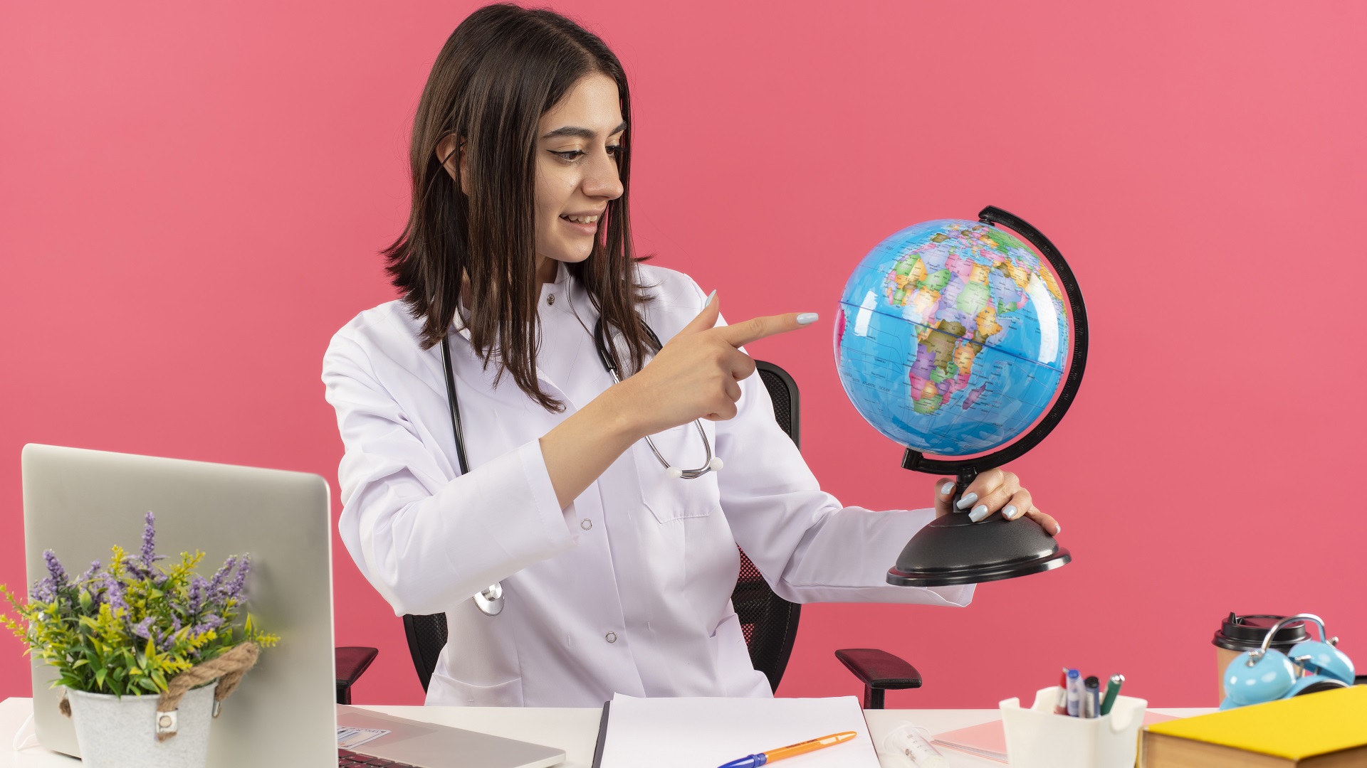 Una mujer sentada frente su escritorio señalando un lugar en un globo terraqueo